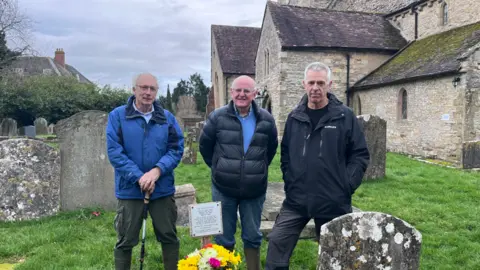 BBC Three men stand in a graveyard and smile at the camera in front of a grave, which has a bouquet of flowers on it.