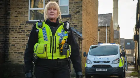 Sarah Lancashire with blonde hair in Ba in her role, where Sergeant Catherine Kawood stands in the police costume and a high green jacket, which contains a radio and a theater connected to it, in front of a lift house from sandstone and a parked police truck in the alley next to it