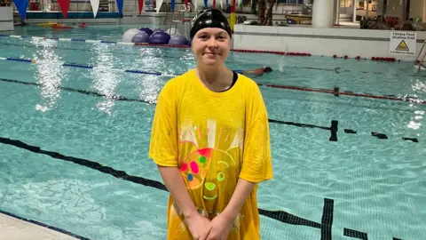 BBC A girl in a big yellow Pudsey shirt and a black swimming cap. There's a pool behind it