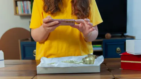 Getty Images A woman with long brown hair wearing a yellow top taking a picture of a gold ornament laying down on a brown table. 