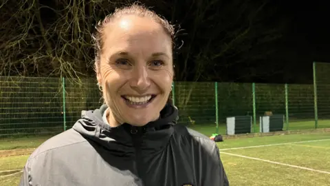 A women is smiling at the camera. She has brown hair tied back in a ponytail. She is wearing a black jacket and is standing on a football pitch. 
