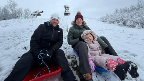 BBC / Jim Scott A man, a woman and a small child are sitting on sledges at the bottom of a hill that is covered in snow. The family are smiling and dressed in winter clothing. In the background, the trees are covered in snow.