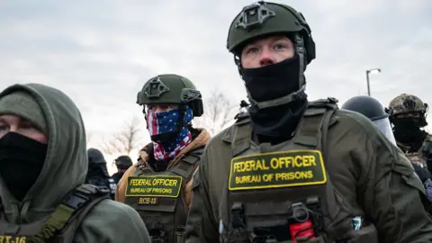 A close up of a federal office in Minneapolis . He is wearing uniform which says 'Federal Officer in yellow accross the front and wears a hemlet and scarf accross his mouth. Another federal office is behiund him with a scarf accross his face with a US flag design on it