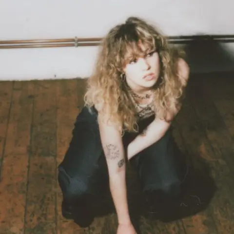 Flower Up A young woman with blonde curly hair and a full fringe crouching on a wooden floor and looking to her left. She has a few small tattoos on her arm and is wearing lots of necklaces and black trousers.