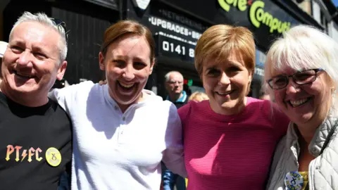 Gambar Ginjal Dari Kiri ke Kanan - Alan Black dengan Rambut Perak dan Kemeja Hitam Membaca Pt FC, Mhairi Black Di Atas Putih, Sturgeon Nicola Di Top Pink dan Ibu Mhairi Black, yang memiliki rambut pirang, mantel abu -abu dan lencana pin SNP SNP pin SNP, lencana SNP pin SNP, SNP pin pin  