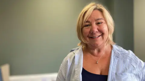 A woman with shoulder-length blonde hair is smiling at the camera in an office. She is wearing an open blue and white striped blouse over a black top.