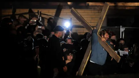 Getty Images It's night. In the right of the photo Michael Sheen is dragging a cross, which is sat on his shoulder. He is clearly uncomfortable. Looking on is someone in black clothes and a black beanie. He gives off a menacing air. In the background is a huge crowd of spectators and media with cameras and lights to be seen. Behind them is the wall of a red-brick community centre. 
