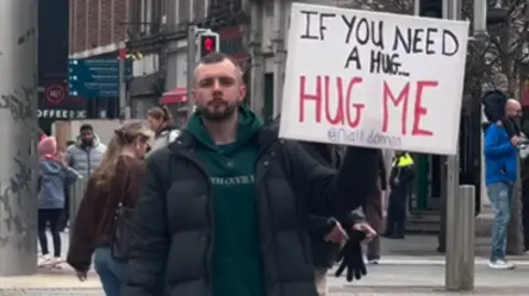 Niall Donnan A man stands in the middle of the footpath in a busy city street wearing a green hoodie underneath a black padded coat. He holds a white sign in red and black writing that reads "If you need a hug... Hug me." 