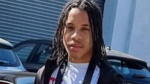 Rhamero West with long brown hair wearing a white t-shirt and black jacket stands in front of two park cars with a business with the shutters down behind him.
