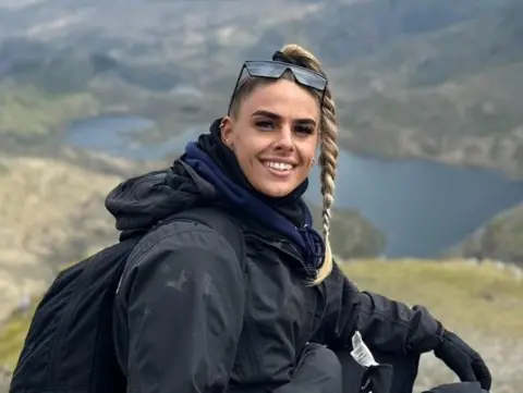 Leonie Hughes A young woman with a blond ponytail, wearing dark walking gear and with sunglasses sitting on top of her head, sits above a view of a lake and mountains