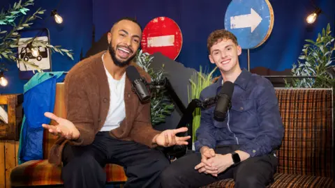 Race Across the World: The Detour Tyler West and Alfie Watts smiling in front of microphones. They are sitting in a recording studio. West is wearing a white T-shirt and brown cardigan and jeans and Watts is wearing a blue jacket and black trousers. 