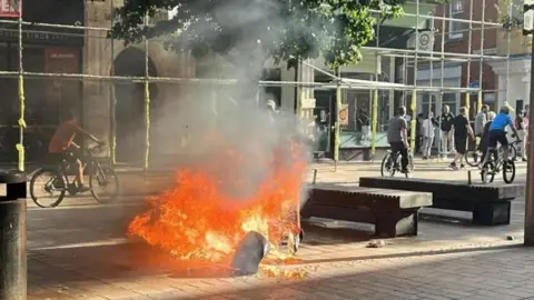 A burning rubbish bin in Hull city centre. In the background, boys ride bicycles towards a small crowd on a street corner.