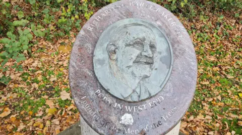 A bronze plaque depicts a man's profile. It sits on top of a stone plinth and on the ground beneath it and behind is green grass covered in brown leaves that have fallen from trees.
