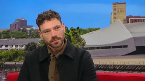 Nelson-Joyce has short, curly brown hair, He wears a blue jacket over a dark green knitted shirt. He has a short dark beard.