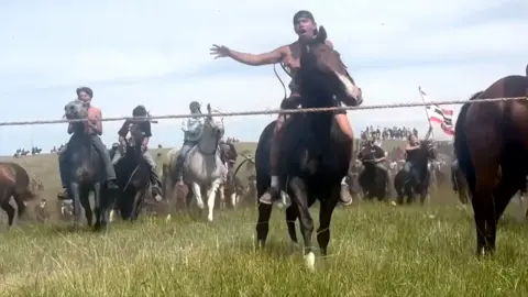 Native Americans on horseback ride down a mound in a field, towards the camera lens, re-enacting the battle of Little Bighorn
