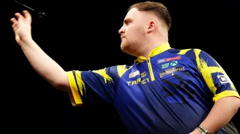 PA Media Luke Littler, with short brown hair and wearing a blue top with yellow edging, throws a dart towards the left of the image.