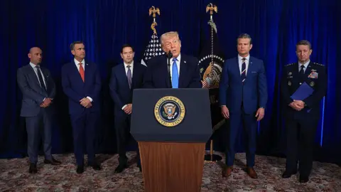 Getty A photograph of Donald Trump standing in the centre at a podium wearing a light blue tie and navy suit, with his team surrounding him.