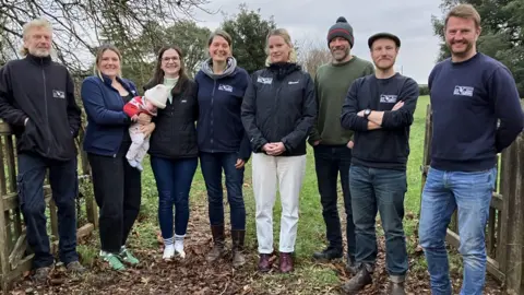 Members of the East Devon District Council Countryside Team stood in a gateway in a field with trees around them. There are eight individuals with one lady holding a baby. 