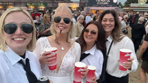 Kym and Tyler Four women enjoying a music festival. Kym is in the middle, wearing a white wedding dress, bald cap and stick-on goatee. Her friends have white shirts and black ties, and two have sunglasses. They are holding red drinks cups.
