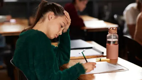 A teenage girl writing on paper while sitting at a desk during exam in classroom. She has one hand on her head and has a concentrated expression. She wears a green jumper and has dark hair pulled back in a ponytail. 