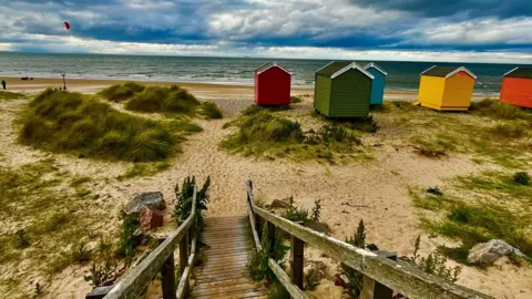 Susan Morrison A wooden walkway or stairway descends into a sandy beach area. Several brightly-coloured beach huts or cabins are visible along the shoreline.