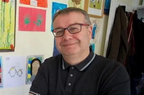 A man stands in a room, with children's pictures on the wall behind him. He has short greying hair and glasses. He is wearing a black polo shirt.
