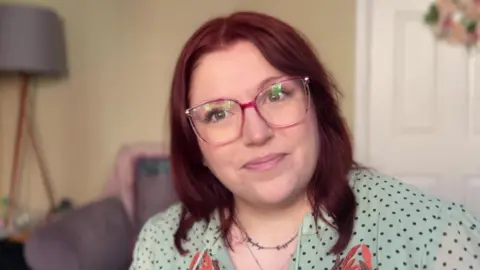 A picture of Hayley, who has dark red hair and glasses and is wearing a green top with black dots on it, there is a door and lamp in the background.