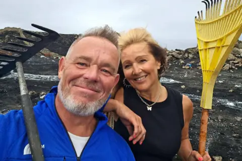 Dean Ward and Allie Evans. He has grey hair and a beard and is wearing a blue top. She has a black top, silver earrings and a silver necklace with butterfly pendant. They are holding rakes and stood on a blackened mountain