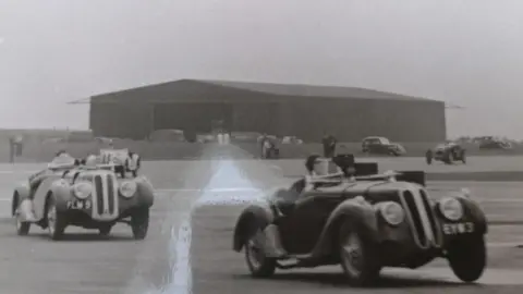 Paul Joslin/BBC A grainy black and white photo showing two cars driving at RAF Gransden Lodge in 1946. There are other vehicles behind them and a handful of spectators. In the distance is a large aircraft hanger. 