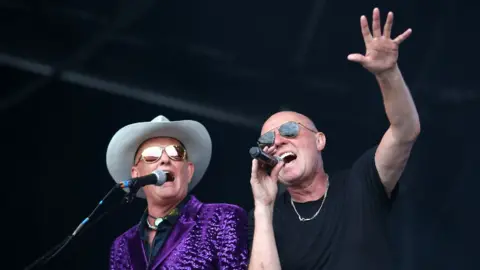 Getty Images Two men on a stage sing into microphones. One wears a black t-shirt and sunglasses and holds his hand up in the air, fingers spread. His band-mate also wears sunglasses, alongside a cowboy hat and purple, sparkly jacket.