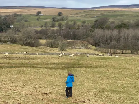 Sara stands in a field with her back to the camera, surveying the landscape beyond. Ahead of her are several, a few copses of trees and green and brown hills and fells in the far distance. She is wearing a thick blue coat and brown hat.