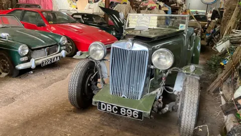 Four classic and vintage cards, all from different eras, are parked in a barn. In the foreground is a dark green car appearing to be from the 1920s or earlier, which has no roof.