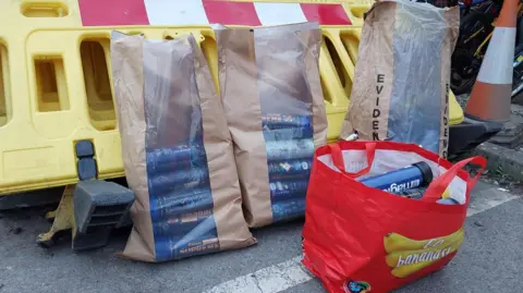 Blue canisters in a brown evidence bag and Aldi shopping bag sitting on the floor, leaning against traffic road signs.
