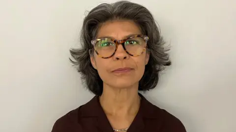 Close-up portrait of Sarah Armstrong wearing round tortoiseshell glasses and a dark collared top, facing the camera against a plain light-colored background.
