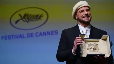 John Travolta holds a sparkling award in box while smiling. He is wearing circular glasses and a white baret with a black suit. In the background is a green and blue screen with the words "Festival De Cannes" written on it. 