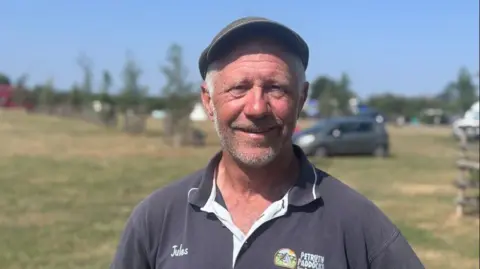 A man is standing with a campsite behind him. He has a navy polo shirt and cap on and is smiling at the camera.