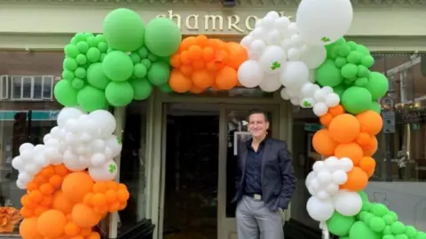 Steven Wells/Shamrock Steven Wells stands in the entrance of his pub. He has short dark hair and wears a navy suit jacket with a navy shirt and grey trousers. Orange, white and green balloons are arranged around the entrance of the pub above and around him.