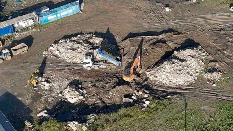 An aerial shot of a lorry tipping waste into piles on a brown patch of earth, with some greenery surrounding it