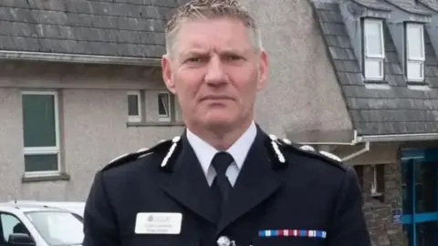 Isle of Man Constabulary Russ Foster looking sternly at the camera. He is wearing his black uniform and name badge and standing in front of police headquarters in Douglas, with is a grey building with a glass sliding door with a blue frame with a car parked outside.