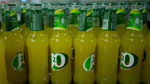 Lots of bottles of apple and mango flavoured J2O on a shelf. The drink is a faint orange in colour and the branding is white and green.