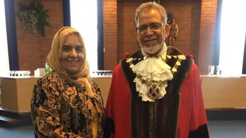 Henna Zia Farhat (left) and Iqbal Zia in a room, both looking at the camera and smiling. Farhat is wearing a cream head scarf and brown and cream patterned jacket. Iqbal is wearing full mayoral robes, with a mayoral chain on, white ruffled shirt, red coat, with brown fur trim. He has glasses on and short grey hair. 