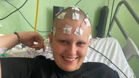 Phoebe Tesoriere Phoebe is sat in a hosptial bed whilst undergoing a diagnostic test. She is smiling with serval medical wires attached to her head which has been shaved.