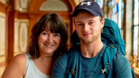 Jo and Sam Gardiner pose together in an ornate room. He is wearing a navy-coloured baseball cap and a blue T-shirt and is carrying a blue rucksack. Jo has shoulder-length dark brown hair and is wearing a light blue vest top. Both are smiling.
