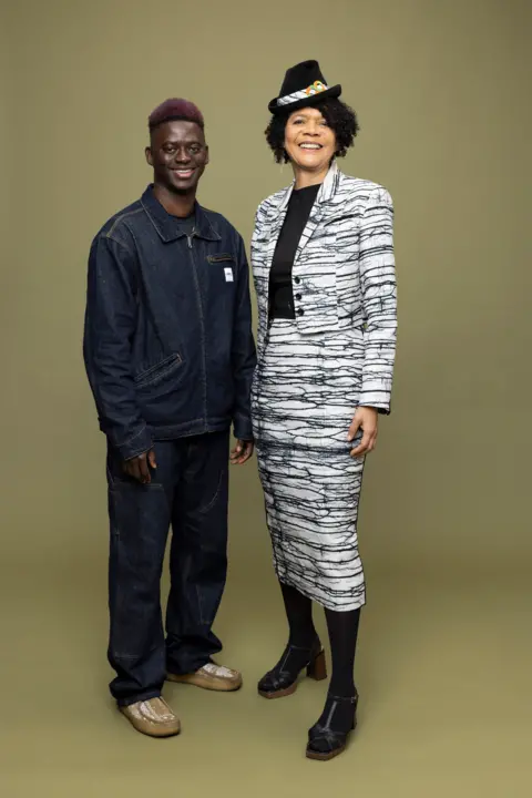 Mark Savage/Newcastle College A vertical image of Chi Onwurah wearing a black and white, almost tiger-print suit jacket over a black top. She is wearing a small black hat with a strip of fabric and two orange and green circles. She is smiling into the camera and standing to the right of Lamin Gibba who has pink hair and is wearing a denim jacket and denim trousers. He is also smiling into the camera.