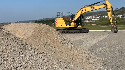 A large pile of rubble in the forefront with grass and houses in the background and a yellow industrial digger in between