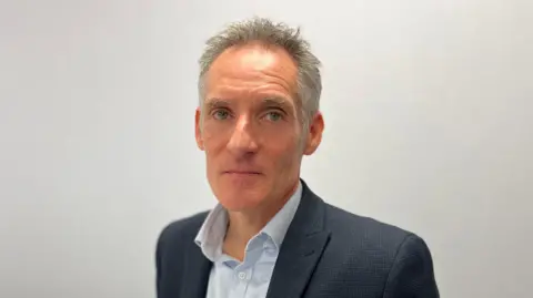 Nick Hynes, the States of Guernsey's director of education, looks at the camera with a neutral expression on his face while stood in a room with a grey wall. He has a navy blue blazer on an a light blue shirt. He has short grey hair.