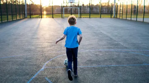 A thin boy wearing a blue T-shirt, dark trousers and trainers dribbles a football in an empty outdoor, fenced basketball/football court. The sun seems to be setting and there is a field outside the court area 