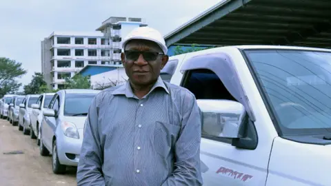 A man wearing a white hat, dark glasses and a shirt stands in front of a line of vehicles.