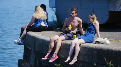 PA Media A man and a women sit on the edge of Bristol Harbour on a sunny day. He is wearing shorts and has his top off, and she is wearing a blue summer dress