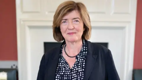 PA Media Sue Gray wearing a navy blouse with achromatic  spots, a small, beaded navy necklace, and a navy overgarment   smiles somewhat  successful  a Downing Street office.